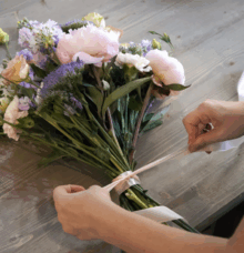Händer binder ett band runt en bukett med blommor på ett träbord.