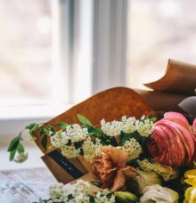 Blommor i olika färger och sorter ligger på ett bord framför ett fönster, inslagna i brunt papper.
