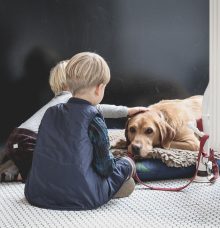 Two children sit on a tiled floor, petting a resting dog lying on a cushion.